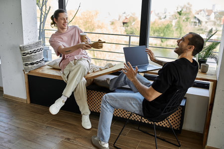 A young gay couple enjoys a coffee break in their modern home, laughing and talking while one uses a...