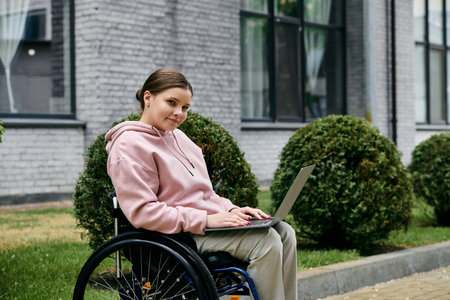 A young woman in a pink hoodie sits in a wheelchair outside, using a laptop.