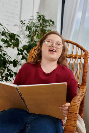 A little girl with Down syndrome laughs while reading a book in a wicker chair.