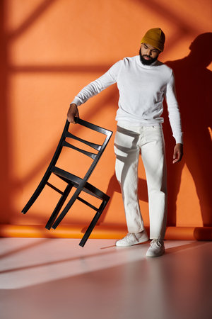 Handsome man in fashionable attire holding chair in front of vibrant orange wall.の写真素材