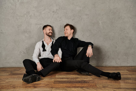 A loving gay couple, dressed in elegant suits, sit together on a wood floor.