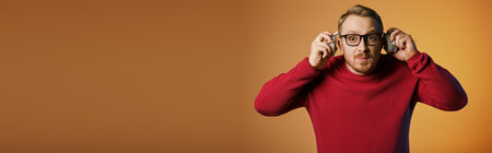 A dapper man in a red sweater gracefully holds a pair of glasses and headphones.