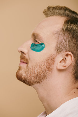 A handsome man in casual attire, sporting a blue eye patch, engaging in a skin care routine.の写真素材