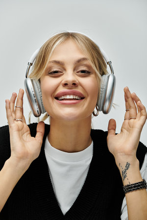 A young blonde woman in stylish casual attire smiles while listening to music through headphones.
