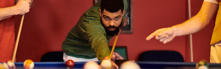 A man in a green sweater lines up a shot at a billiard table while his friends watch, enjoying a...