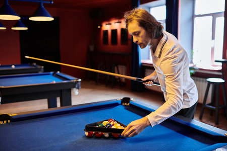 A man in a casual white shirt sets up a game of billiards in a dimly lit pool hall.