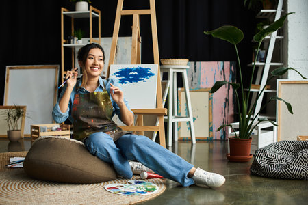 A young Asian artist takes a break from painting, sitting on a beanbag in her art-filled studio.の写真素材