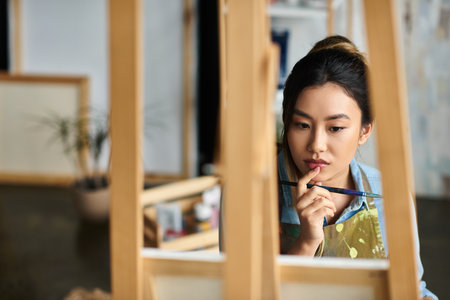 A young Asian artist, wearing an apron, thoughtfully considers her next brushstroke in her workshop.の写真素材