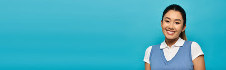 A young Asian woman wearing a white shirt and a blue vest smiles for the camera in front of a light...