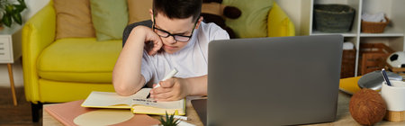 Boy with Down syndrome writing in notebook at table, looking at laptop.の写真素材