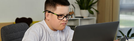 A young boy with Down syndrome sits at a desk, intently looking at his laptop.