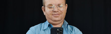 A man with inclusivity wearing glasses looks at his smartphone in front of a black background.