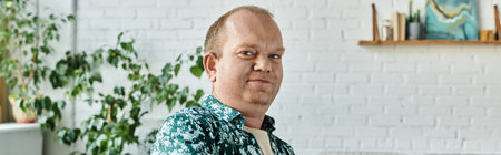 A man with inclusivity in a patterned shirt stands against a white brick wall.