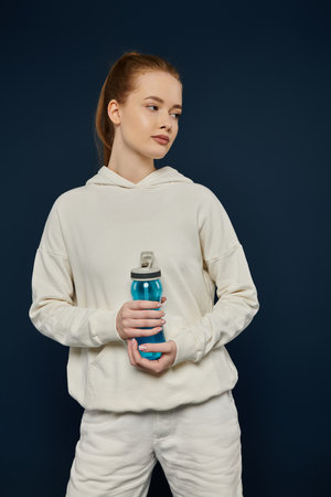 Young woman in white hoodie and pants poses with water bottle against blue backdrop.