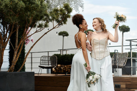 Two brides stand on a rooftop terrace, smiling and holding bouquets.の写真素材