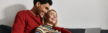 A happy multicultural couple shares a laugh together on a comfortable sofa in their modern...