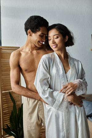 An Asian woman in white robe embraces an African American man in a bedroom, close with faces inches apart, showing intimacy.の写真素材