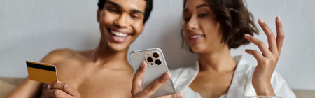 A young, multicultural couple shares a laugh in their bedroom, the man holding a credit card and the woman a phone.の写真素材