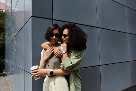 Two women in a loving embrace stand against a modern office building.の写真素材