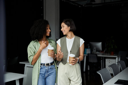 Two women in an office setting, holding coffee cups and smiling at each other.の写真素材