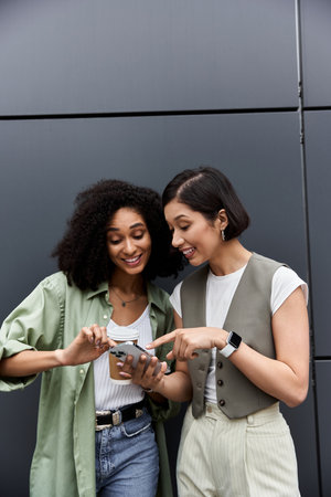 Two women in casual attire stand outside, looking at a phone.の写真素材