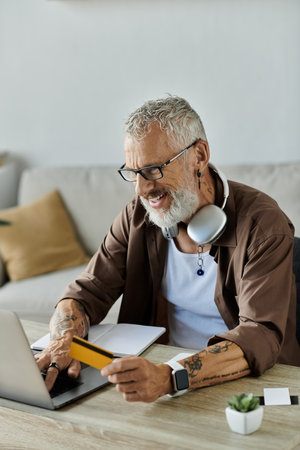 A mature gay man with grey hair and tattoos works remotely from home, using a laptop and credit card, with a smile.の写真素材