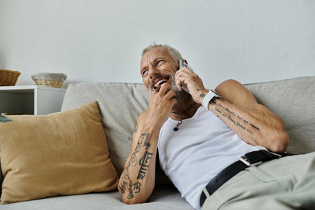 A mature gay man with tattoos and a grey beard sits on a couch at home, laughing as he talks on the...