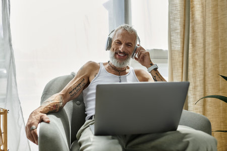 A mature gay man with tattoos and a grey beard relaxes on a couch at home while wearing headphones...