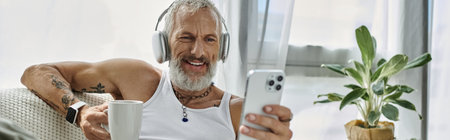 A mature man with grey beard and tattoos is smiling and relaxing on a couch at home. He is wearing...
