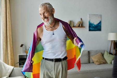 A mature gay man with tattoos and grey hair smiles while holding a rainbow pride flag in his home.の写真素材