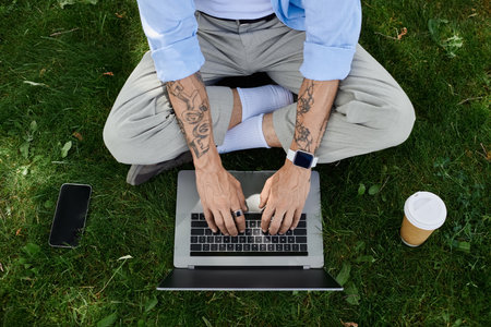 A mature man with tattoos sits on the grass typing on his laptop outdoors.
