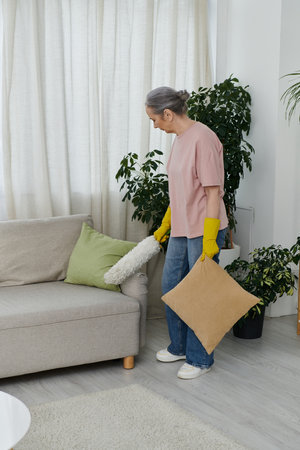 A mature woman dusts the couch in her modern apartment.