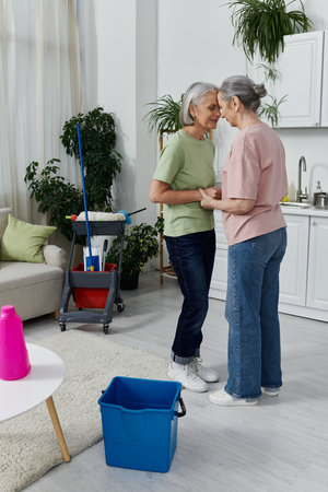 Two mature women, a lesbian couple, take a break from cleaning their modern apartment. They are standing in the living room, holding hands.の写真素材