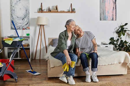 Two mature lesbian women relax on their bed in a modern apartment after cleaning, showcasing a loving relationship and a comfortable home.の写真素材