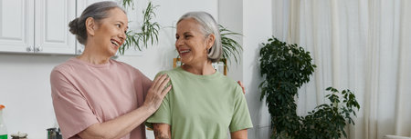 Two older lesbians laugh in modern apartment, one finishes housework.