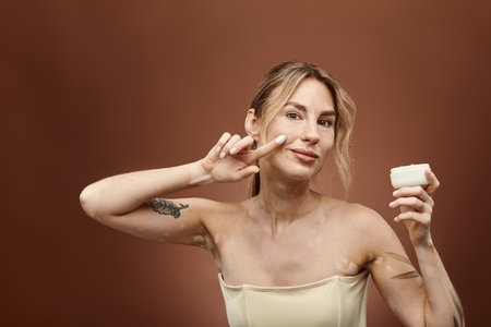 Young woman with vitiligo smiles, applying cream to her face and holding a jar, looking directly at the camera.の写真素材