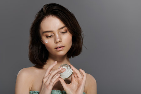 A young woman with short dark hair applies face cream against a gray backdrop.