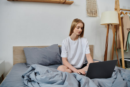 A young woman sits on a bed, working on a laptop.の写真素材