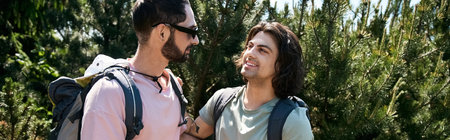 A young gay couple smiles and walks together in a sunny forest during a summer hike.