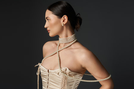 A young woman with brunette hair wears a pearl necklace and looks to the side against a dark...