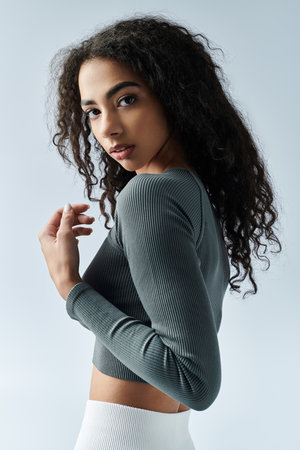 A young Black woman with curly hair, wearing a long-sleeved shirt, stands confidently on a white...