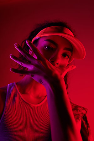 A young woman in a tank top and visor poses dramatically against a vibrant red backdrop illuminated by neon lights.の写真素材