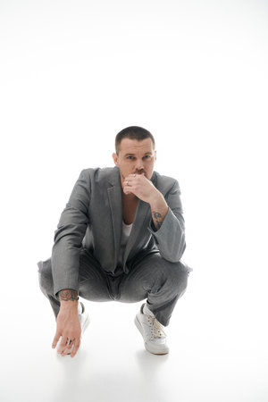 A man in a gray suit and white sneakers sits in a squat position.