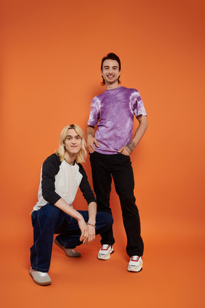 Two young queer friends strike a pose in colorful, stylish attire against an orange backdrop.