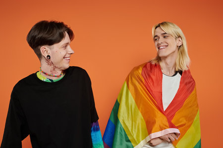 Two young queer people with pride rainbow flag against an orange background.