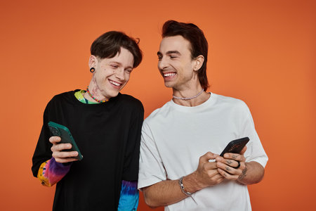 Two young queer friends in vibrant outfits laugh while looking at their phones against an orange...