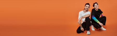 Two young friends, dressed in stylish attire, smile at the camera while sitting against an orange background.の写真素材