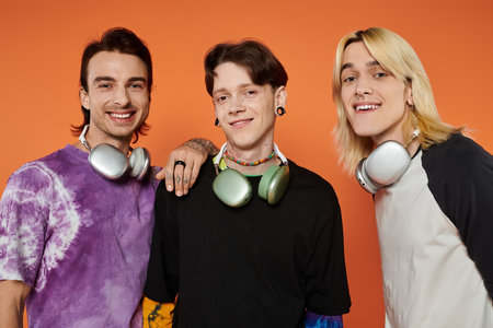 Three young friends, wearing stylish attire, pose with headphones against a vibrant orange background.の写真素材