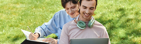 Two young queer friends sit on the grass, one working on a laptop, the other writing on paper.