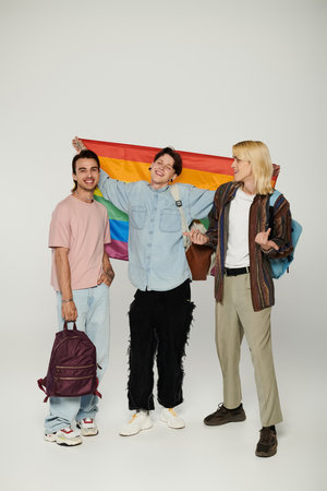 Three young students, part of the LGBTQIA community, stand together holding a rainbow flag.